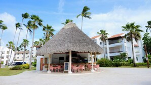A thatched-roof outdoor seating area with tables and chairs, surrounded by palm trees and a white building in the background. - Just  few steps from Beach, Enjoy a Sunrise in this Wonderful place (Punta Cana)
