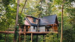 A treehouse with a sloped roof and a wooden deck, surrounded by dense forest. - The Treehouse @Brandywine Grove (Sugarcreek)