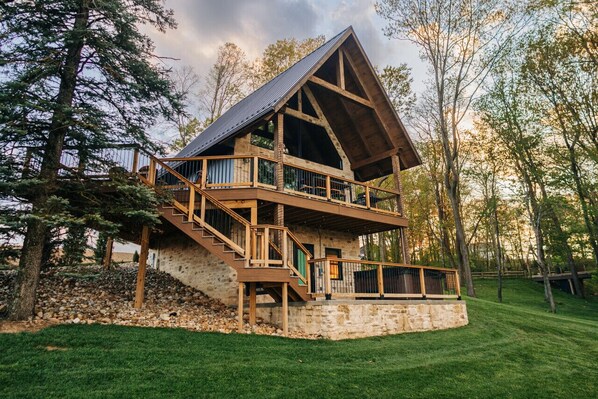 A multi-level wooden cabin with a stone foundation, surrounded by trees and a grassy area.