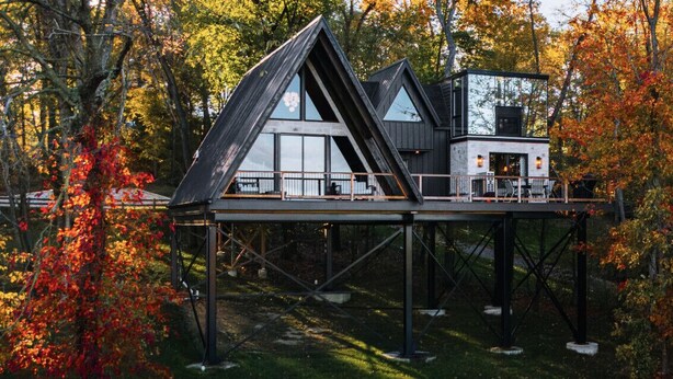 Une maison moderne dotée d’une toiture triangulaire, surélevée sur des piliers, et entourée du feuillage automnal.