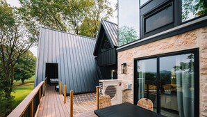 Une terrasse extérieure moderne, dotée d’une table et de chaises en bois, d’une structure en métal et d’un bâtiment en pierre avec de grandes fenêtres.