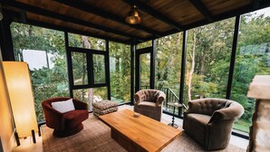 Une pièce spacieuse avec de grandes fenêtres donnant sur une forêt. On y trouve une table basse en bois et deux fauteuils.