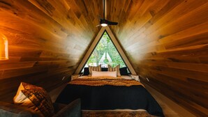 Une pièce triangulaire comprenant un lit, un ventilateur de plafond et une fenêtre donnant sur des arbres.