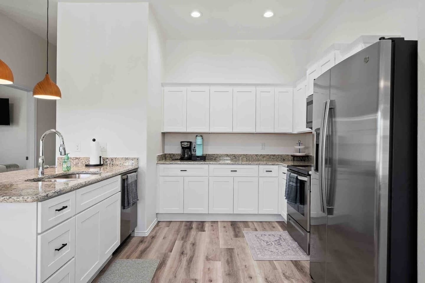 Una cocina moderna con armarios blancos, encimeras de granito, un refrigerador de acero inoxidable y una cafetera.