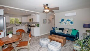A spacious living room with a dark blue sofa, wooden coffee table, and ottomans, a dining area with a wooden table and chairs, and a kitchen with white cabinets and a ceiling fan. - Fallbrook's Finest Hideaway Casa - Pool, Game Rm (Fallbrook)