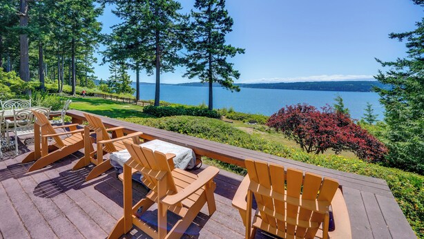 A deck with wooden chairs and a table, overlooking a lake with trees and a distant mountain range. - Cozy Langley Retreat: Water Views & Beach Access (Langley)