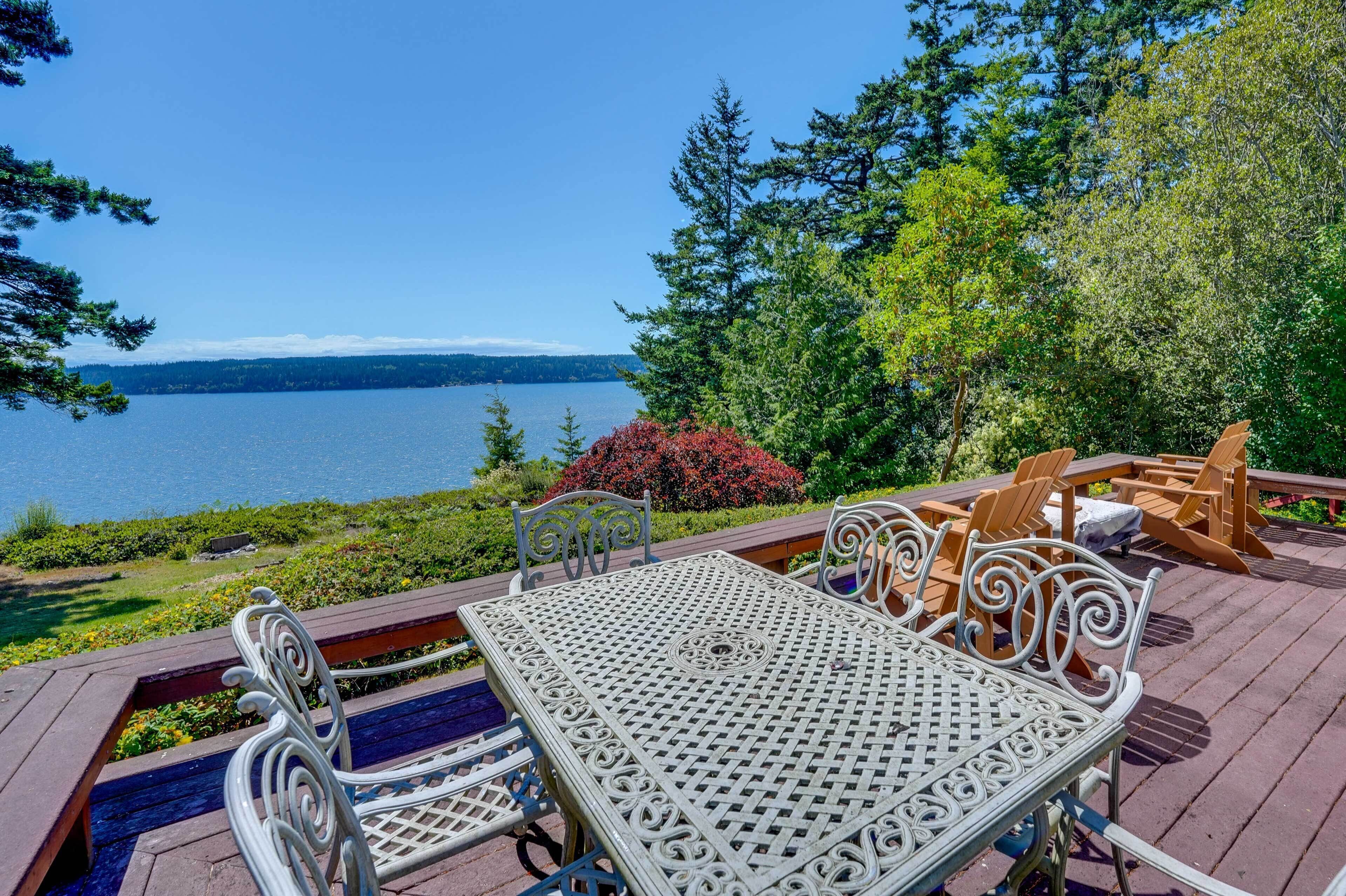 Une terrasse avec une table et des chaises, donnant sur un lac bordé d’arbres et d’arbustes.