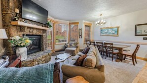 A cozy living room with a stone fireplace, a flat-screen TV, a dining area with a wooden table and chairs, and a large window with blinds.