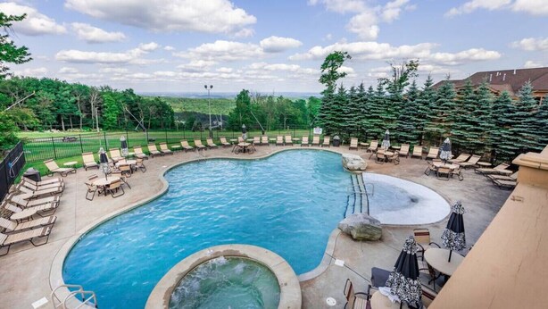 A swimming pool with lounge chairs and a hot tub, surrounded by trees and a view of the landscape.