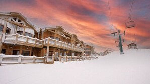 A wooden chalet-style building with a balcony, surrounded by snow, and a ski lift in the background.