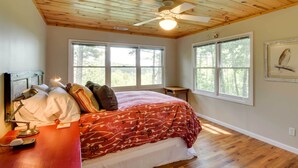 A bedroom with a wooden ceiling, a bed with a red floral bedspread, a desk with a lamp, and a framed bird picture on the wall. - Spacious Georgia Escape w/ Fireplace, Deck & Grill (Lakemont)