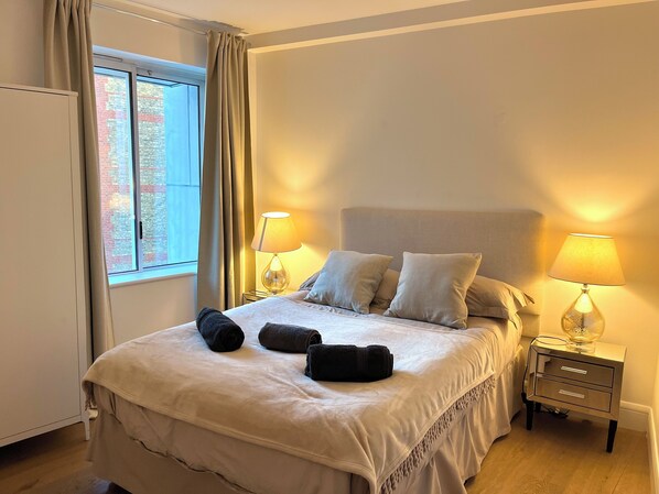 Ein ordentlich bezogenes Bett mit zwei Kissen, ein Nachttisch mit einer Lampe und ein Fenster mit Vorhängen.