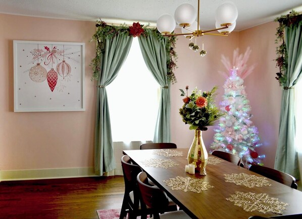 Un comedor con una mesa de madera dispuesta para cuatro, un florero con flores, un árbol de Navidad con luces y una obra de arte enmarcada en la pared.