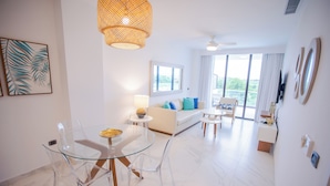 Una sala de estar moderna con una mesa de comedor de vidrio, sillas traslúcidas, un sofá blanco con cojines azules, una mesita auxiliar con lámpara y un ventanal con cortinas.