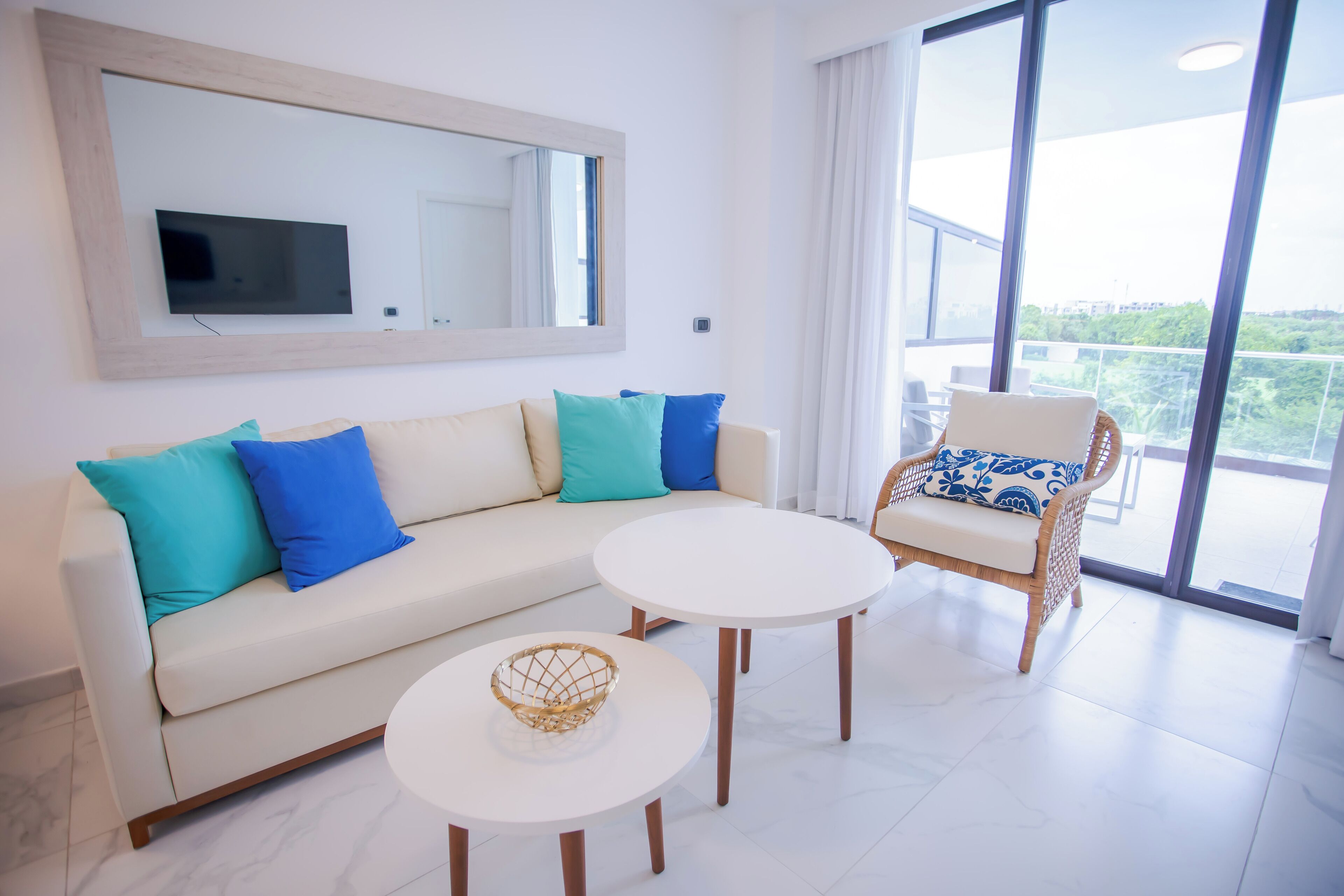 Una sala de estar moderna con un sofá, dos mesas redondas, un sillón de mimbre, un espejo grande y un televisor.