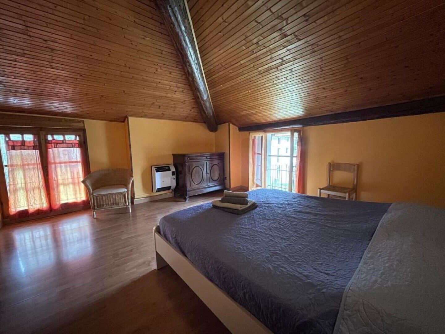 A bedroom with a wooden ceiling, a bed, a chair, a cabinet, and a heater.
