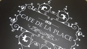 Ein schwarzer Tisch mit weißen dekorativen Rändern und der Aufschrift "CAFE DE LA PLACE 1970" in der Mitte.