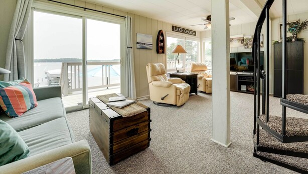 A spacious living room with a sofa, armchair, and a wooden chest of drawers. A sign reading "LAKE GEORGE" is visible on the wall.