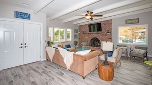 A spacious living room with a fireplace, a ceiling fan, and a large window overlooking a pool. - Fallbrook's Finest Hideaway House - Pool, Game Rm (Fallbrook)