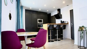A modern kitchen with a dining area, featuring a round table with chairs, a bar with stools, and a black TV mounted on the wall.