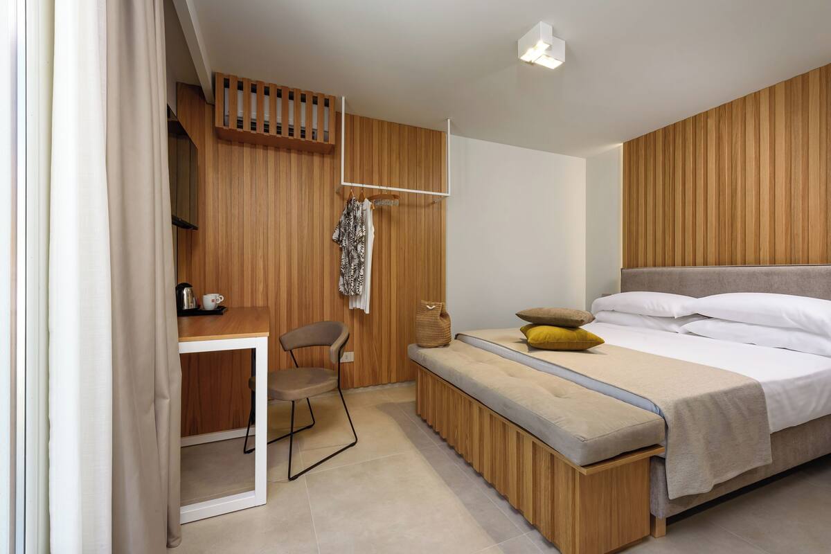 A bedroom with a wooden slatted wall, a bed with pillows, a bench, a chair, a small table with a cup, and a hanging coat rack.