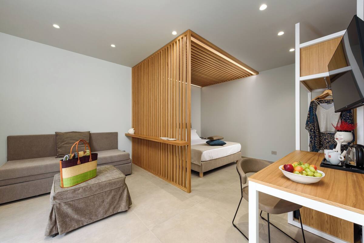 A modern, compact room with a sofa, a bed, a wooden partition, a dining table with a bowl of fruit, and a hanging coat rack.