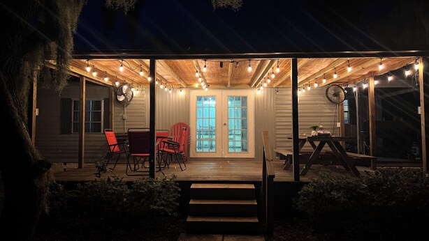 Une terrasse extérieure couverte, agrémentée de guirlandes lumineuses, d’une plateforme en bois et d’un coin repas avec des chaises rouges.