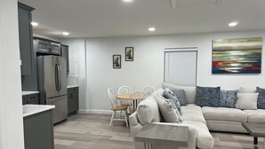 Une cuisine moderne avec un réfrigérateur en acier inoxydable, un coin repas avec une table en bois et des chaises blanches, et un salon avec un canapé gris et une table basse.