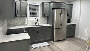 Une cuisine moderne avec des meubles gris, un réfrigérateur en acier inoxydable, un évier avec un robinet noir et une fenêtre équipée de stores.