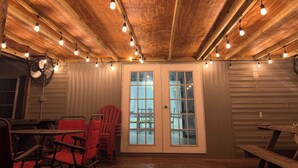 Un intérieur chaleureux avec des poutres en bois au plafond, des guirlandes lumineuses, un ventilateur et une vue à travers une porte vitrée.