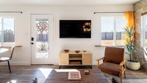 A living room with a wooden floor, a brown leather chair, a TV on the wall, a door with glass panels, and a potted plant. - New! The Aberdeen House - Private Acreage + Joshua Trees + Hot Tub! (Yucca Valley)