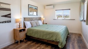 A bedroom with a wooden bed, two bedside tables with lamps, a wall-mounted air conditioner, a window with a view of the desert, and a framed picture on the wall. - New! The Aberdeen House - Private Acreage + Joshua Trees + Hot Tub! (Yucca Valley)