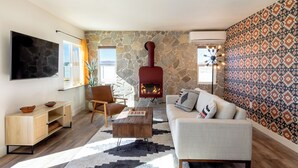 A living room with a stone wall, a fireplace, a sofa, a coffee table, and a TV mounted on the wall. - New! The Aberdeen House - Private Acreage + Joshua Trees + Hot Tub! (Yucca Valley)