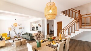 Uma sala de estar ampla, com uma mesa de jantar de madeira posta para quatro pessoas, rodeada por cadeiras brancas. Uma escada de madeira com corrimão leva a um andar superior.