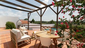 Um pátio externo com mesa e cadeiras de madeira, um sofá branco e uma piscina.