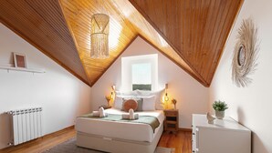 Quarto com teto inclinado, cama com travesseiros e cobertor, mesas de cabeceira com abajures, radiador e um pequeno armário.