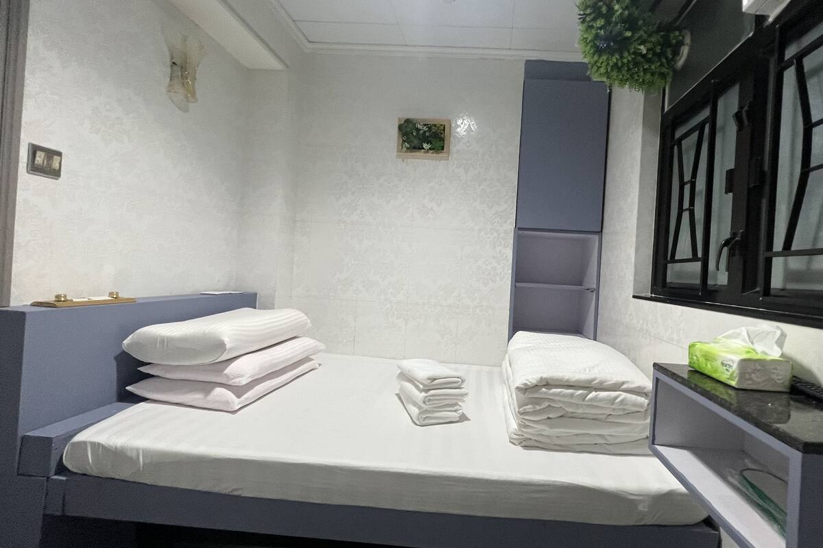 A neatly made bed with folded white linens, a small bedside table with a tissue box, and a window with black frames.