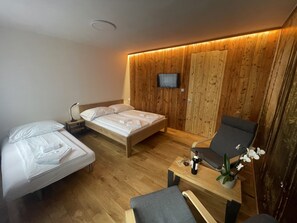 A modern hotel room with a wooden wall panel, a bed, a desk with a chair, a TV mounted on the wall, and a small table with a bottle and glass.