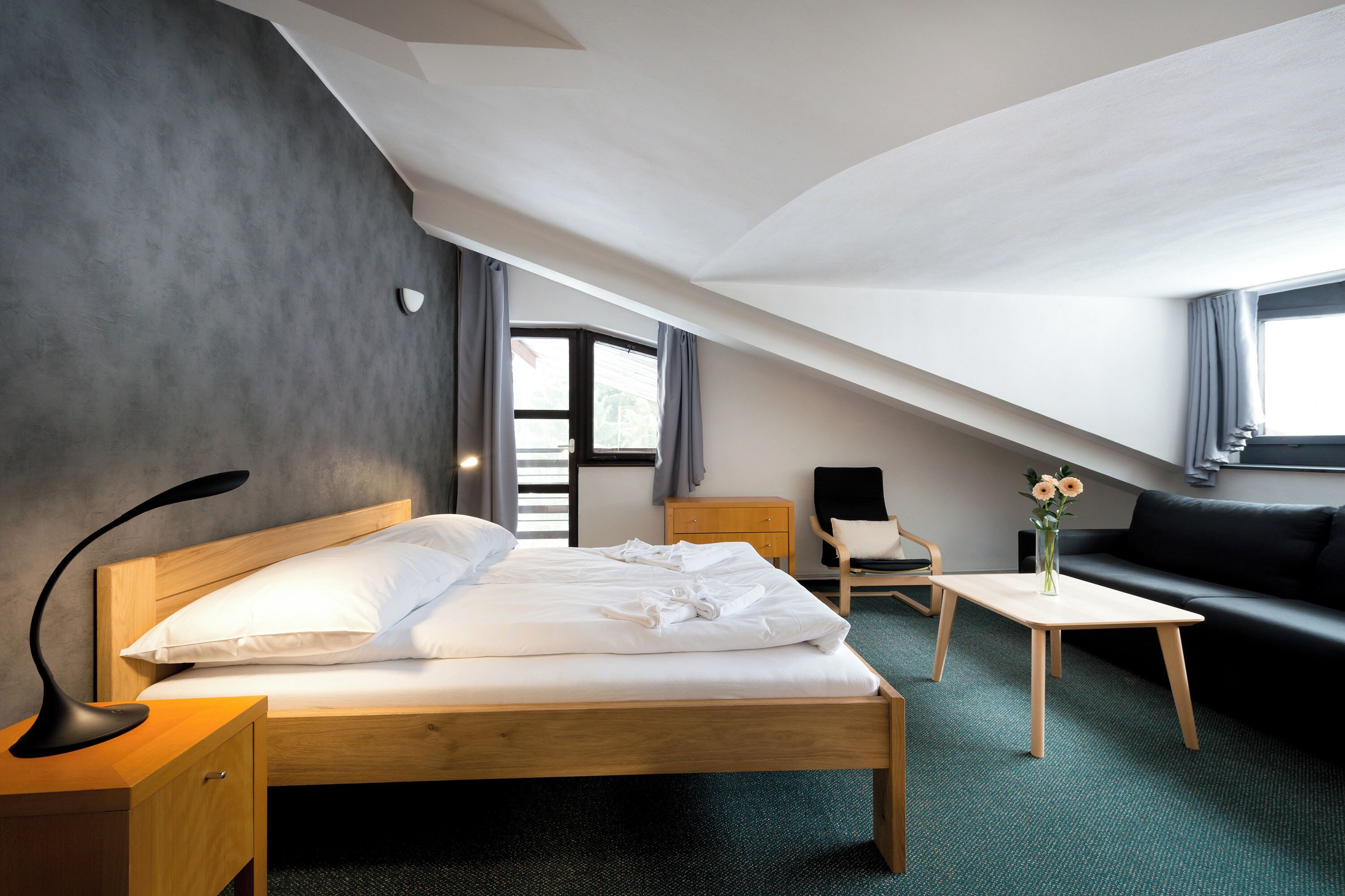 A modern hotel room with a wooden bed, a small desk, a sofa, and a view of the outdoors through a window.
