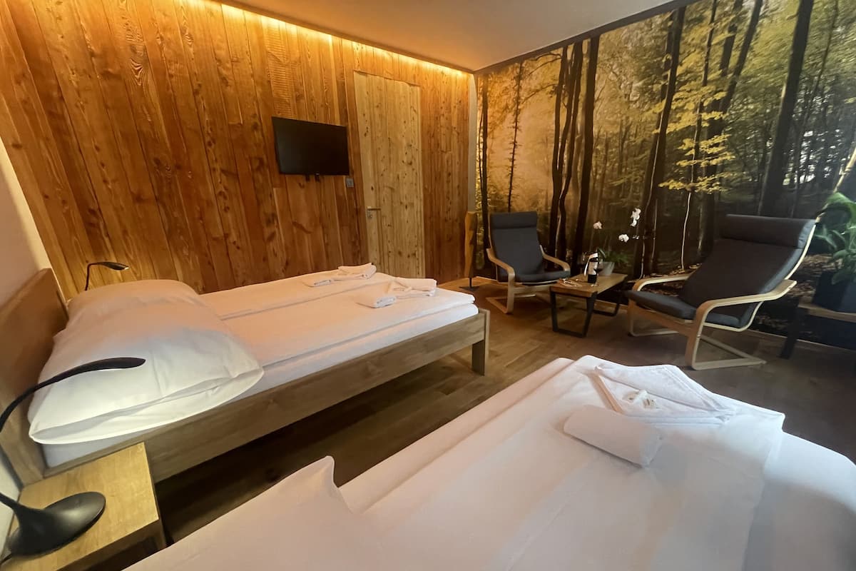 Une pièce avec un mur en bois, un lit recouvert de draps blancs, deux chaises, une petite table et un téléviseur fixé au mur.