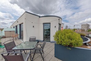 Un bâtiment moderne à la façade incurvée, doté d’un espace extérieur avec des chaises et une table, et d’un grand arbuste au premier plan.