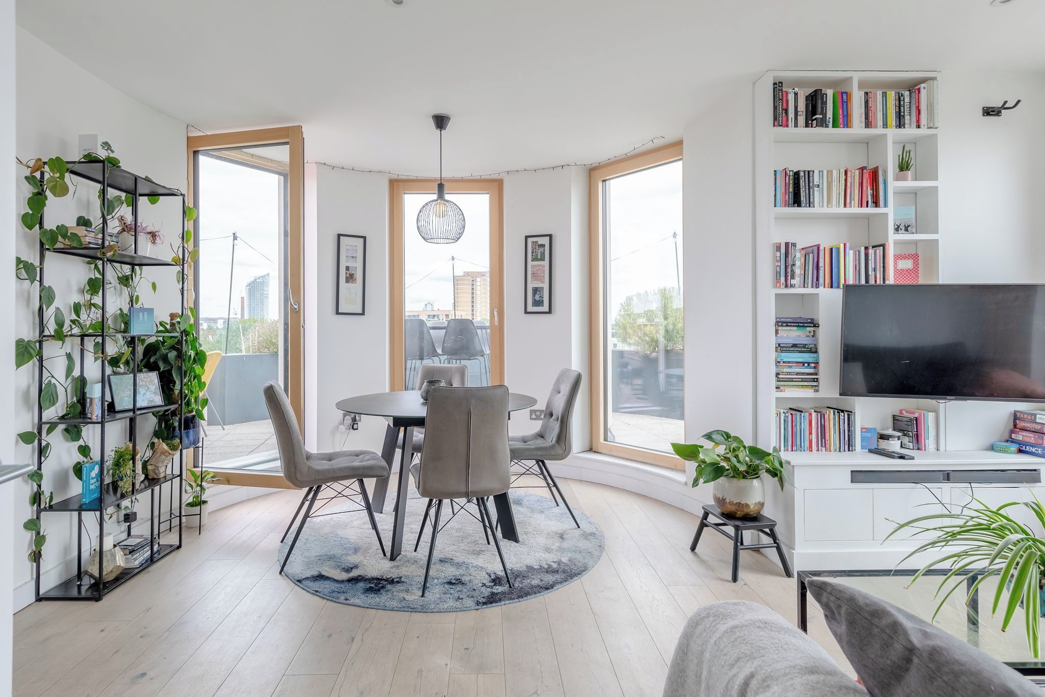 A modern living room with a dining area, large windows, a bookshelf, and a TV.