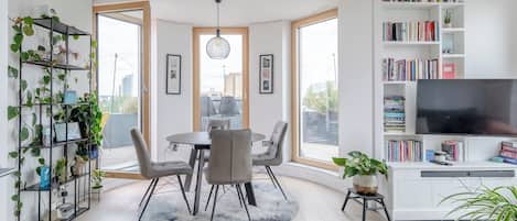 A modern living room with a dining area, large windows, a bookshelf, and a TV.
