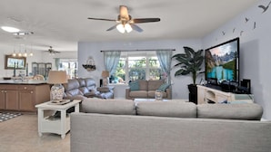 A spacious living room with a large sofa, a dining area with a chandelier, and a TV displaying a scenic landscape. - South Gulf Cove 4BR/2BA Heated Pool ,15 mins to the GULF (Port Charlotte)
