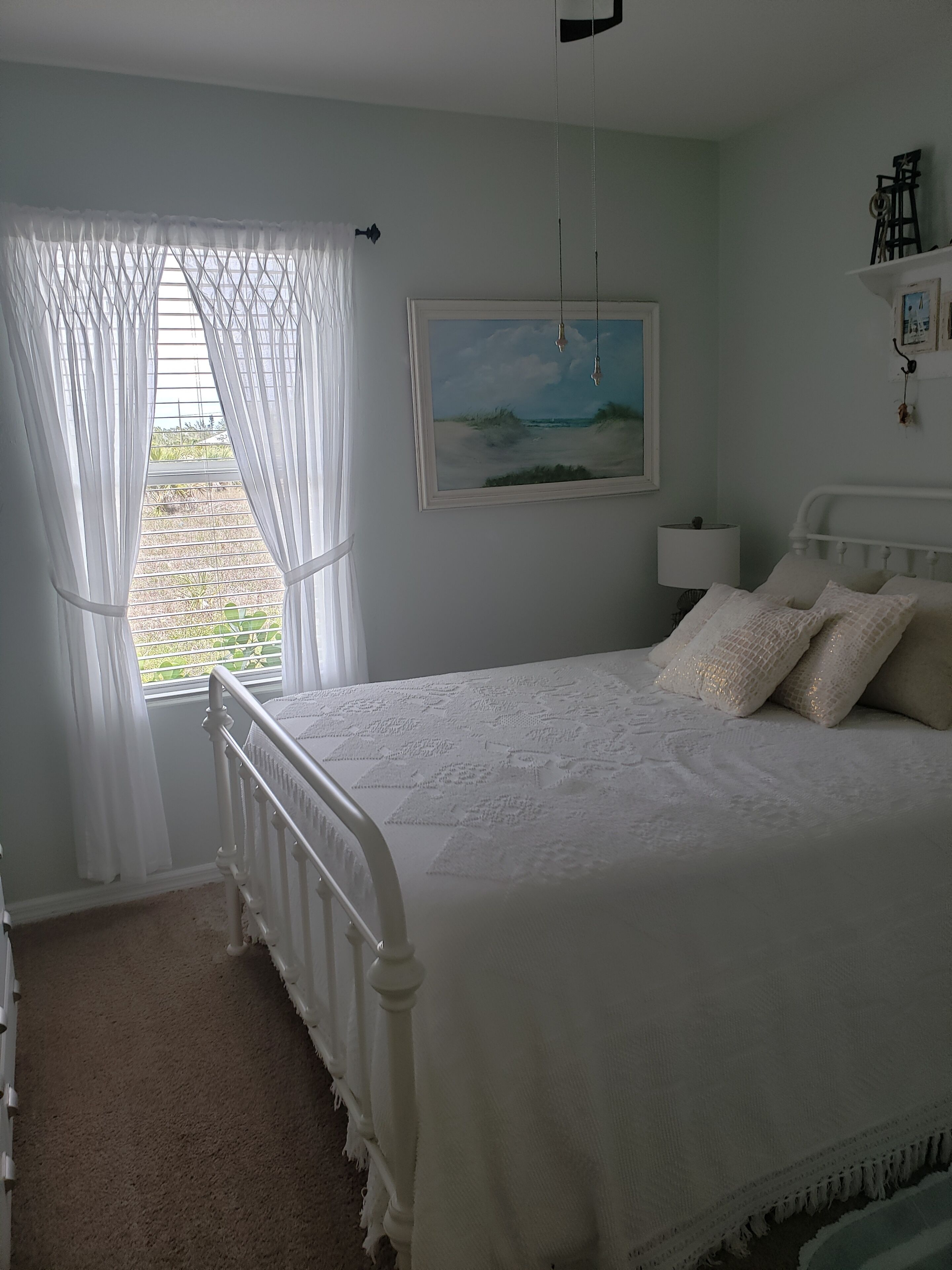A bedroom with a bed, a window with sheer curtains, a painting on the wall, and a bedside table with a lamp.