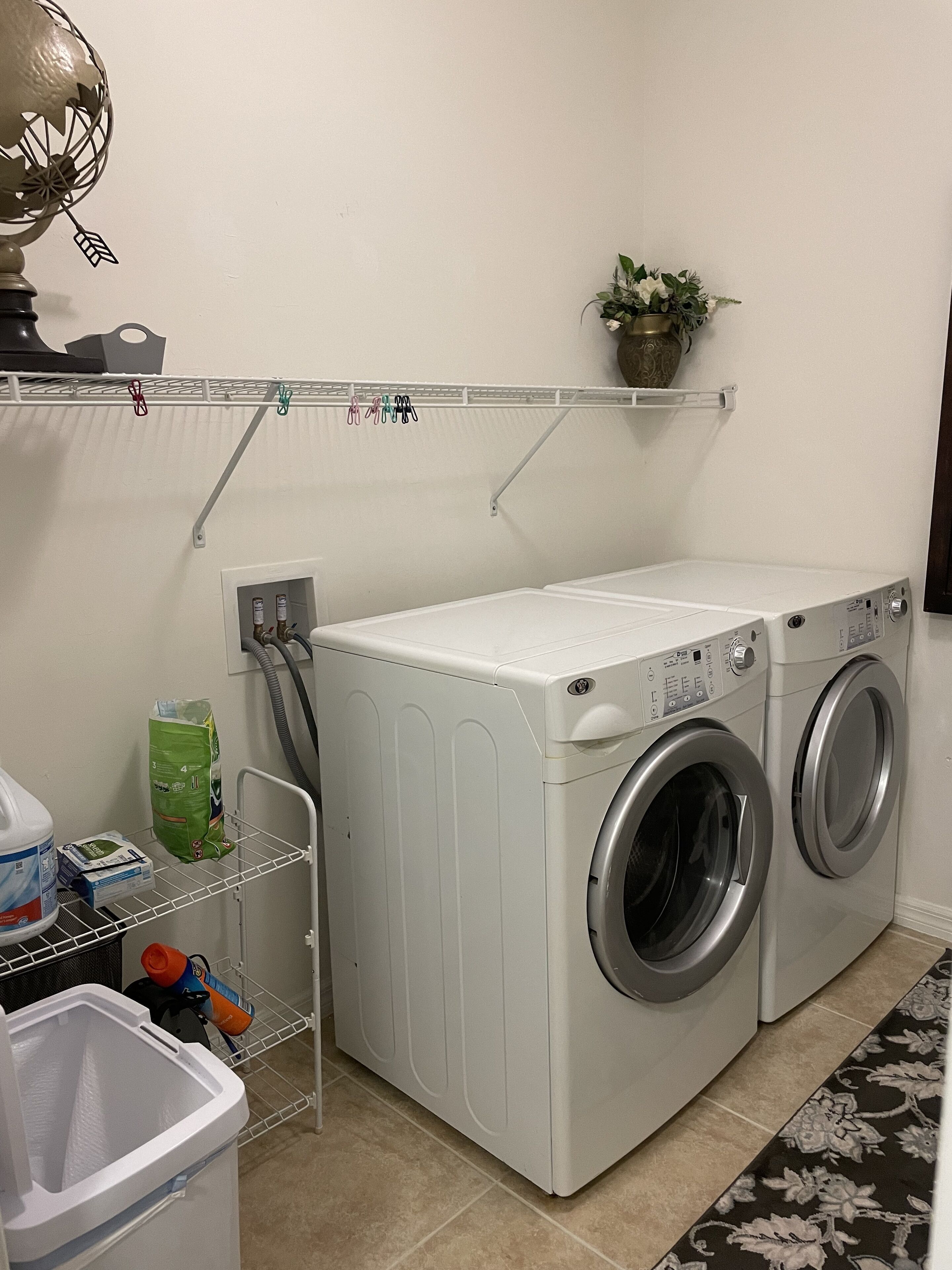 A laundry area with a front-loading washer and dryer, a shelf with cleaning supplies, and a potted plant on the wall.