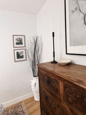 Una habitación con un aparador de madera, un florero blanco con ramas, dos cuadros enmarcados, una vela y una alfombra.