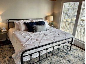 A neatly made bed with a quilted white comforter and several pillows, a bedside table with a lamp, and a window with blinds.
