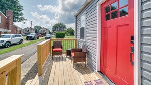 A wooden deck with wicker furniture, a red door, and a view of parked cars and houses in the background.