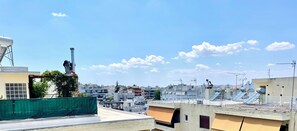 A rooftop with a water tank, a green fence, and a view of a cityscape with buildings and a clear sky.
